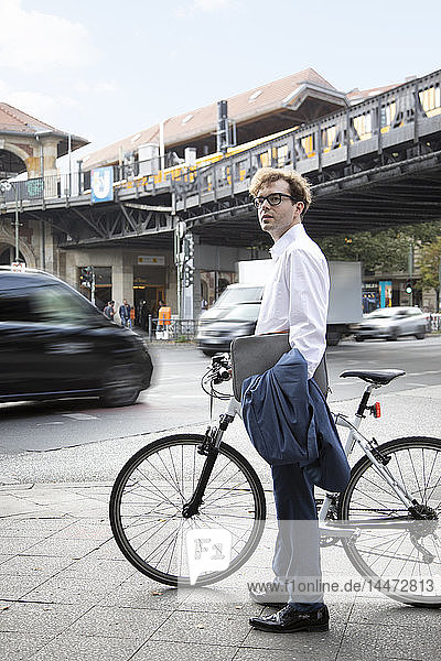 Deutschland  Berlin  Porträt eines auf dem Bürgersteig stehenden Geschäftsmannes mit Fahrrad und Laptoptasche