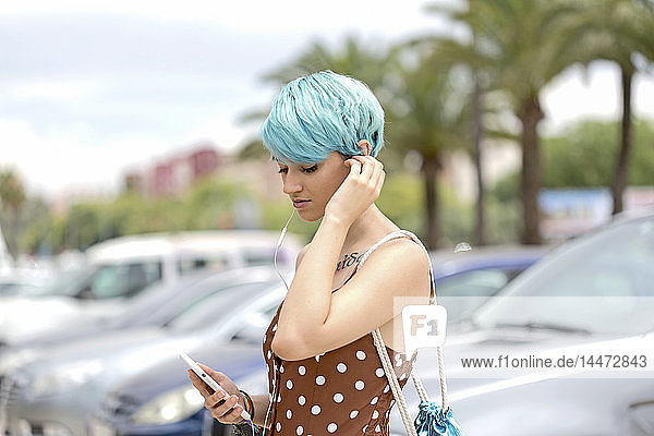 Spanien  junge Frau mit blau gefärbten Haaren  die mit Kopfhörern und Smartphone Musik hört