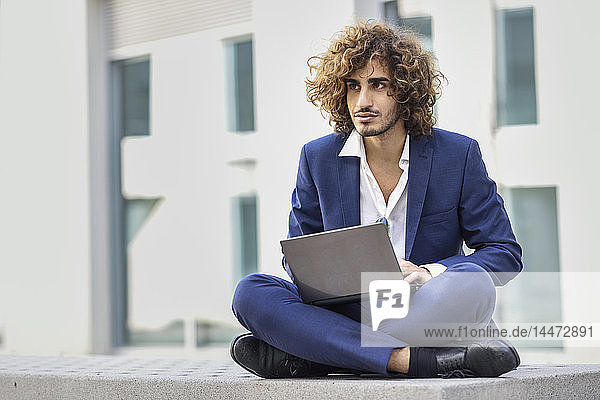 Portrait of young businessman with curly hair wearing blue suit sitting on bench using laptop outdoors