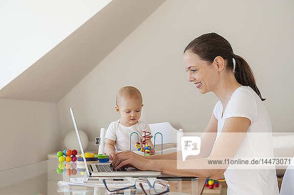 Lächelnde Mutter benutzt Laptop und kleine Tochter spielt zu Hause am Tisch