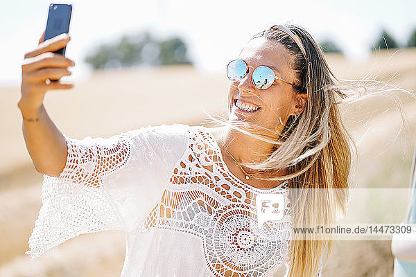 Porträt einer blonden jungen Frau mit verspiegelter Sonnenbrille  die mit einem Smartphone Selbstgespräche führt