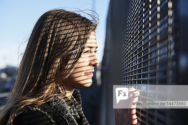 Porträt einer jungen Frau mit Schatten im Gesicht  die durch einen Metallzaun blickt