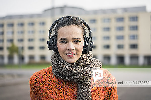 Porträt einer lächelnden Frau mit Schal und orangem Strickpullover  die mit Kopfhörern Musik hört