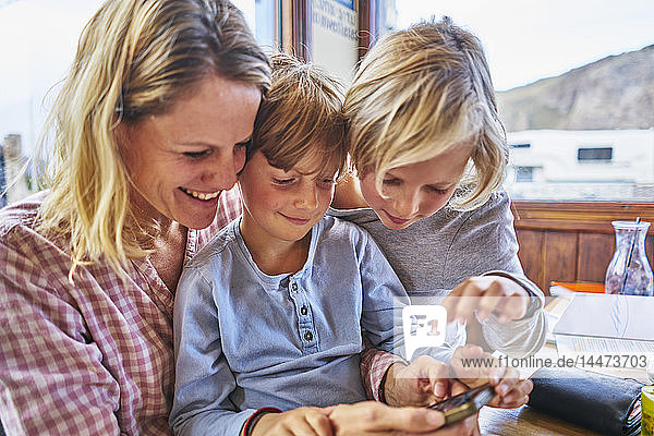 Glückliche Mutter mit zwei Söhnen  die in einem Café am Tisch sitzen und mit dem Handy spielen
