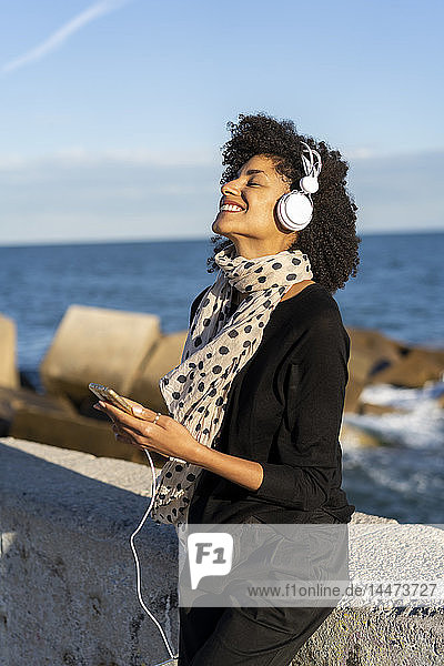 Lächelnde Frau beim Musikhören mit Smartphone und Kopfhörern am Meer
