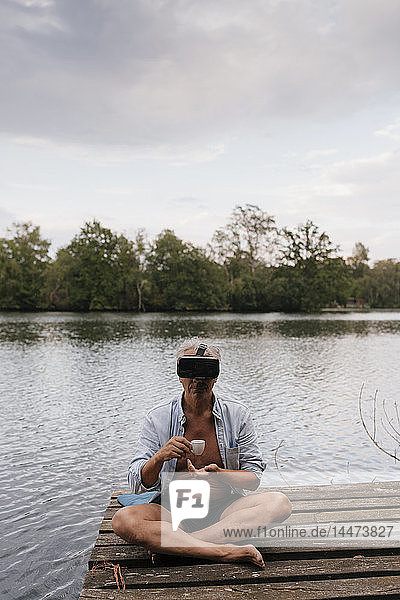 Älterer Mann sitzt auf einem Steg an einem See  trägt eine VR-Brille und trinkt Kaffee