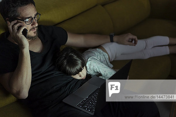 Father talking on cell phone on couch at night with daughter lying next to him