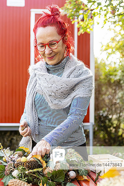 Porträt einer lächelnden älteren Frau mit rot gefärbten Haaren beim Basteln an der Herbstdekoration im Garten