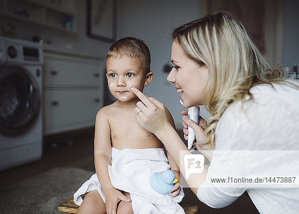 Lächelnde Mutter trägt zu Hause im Badezimmer Hautcreme auf das Gesicht des Kleinkindes auf