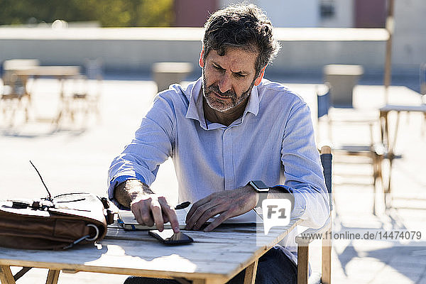 Reifer Mann sitzt am Tisch im Freien mit Handy und Notebook