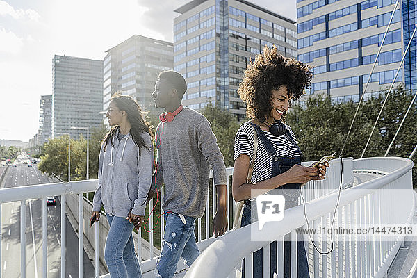 Junge Frau mit Kopfhörern steht auf der Brücke  benutzt ein Smartphone  junge Leute gehen vorbei