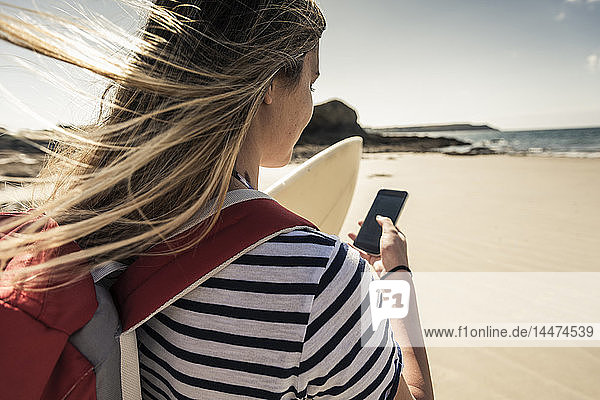 Junge Frau am Strand  mit Surfbrett  mit Smartphone