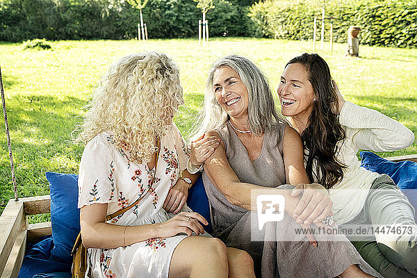 Drei glückliche Frauen unterschiedlichen Alters sitzen auf einem hängenden Bett im Garten und unterhalten