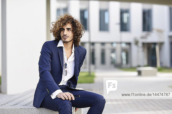 Portrait of young businessman with curly hair wearing blue suit sitting on bench outdoors