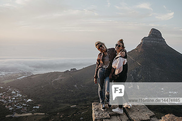 Südafrika  Kapstadt  Kloof Nek  Porträt von zwei glücklichen Frauen  die bei Sonnenuntergang auf einem Felsen stehen