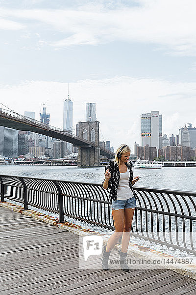 USA  New York City  Brooklyn  junge Frau steht mit Kopfhörern und Handy am Wasser