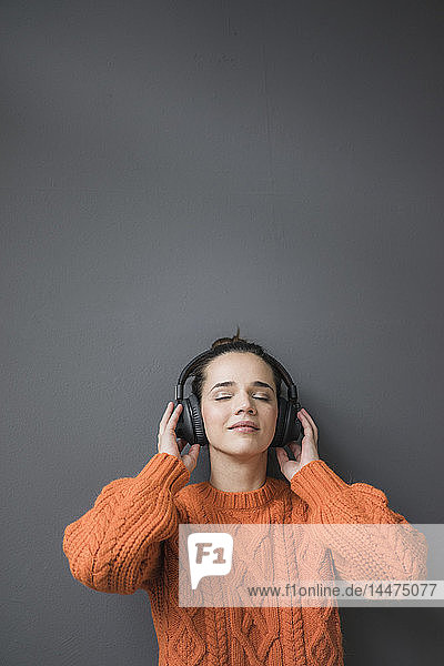 Porträt einer Frau in einem orangefarbenen Strickpullover  die sich an eine graue Wand lehnt und mit Kopfhörern Musik hört