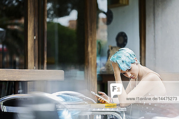 Nachdenkliche junge Frau mit blau gefärbten Haaren sitzt in einem Straßencafé und hört Musik mit Kopfhörern