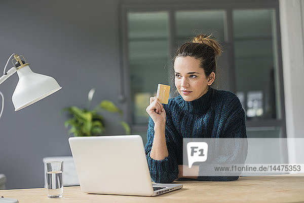 Porträt einer selbstbewussten Frau mit Laptop und Kreditkarte  die zu Hause am Schreibtisch sitzt und in die Ferne schaut