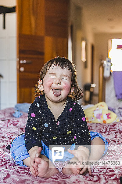 Porträt eines barfuss auf dem Bett sitzenden Kleinkindes mit herausgestreckter Zunge