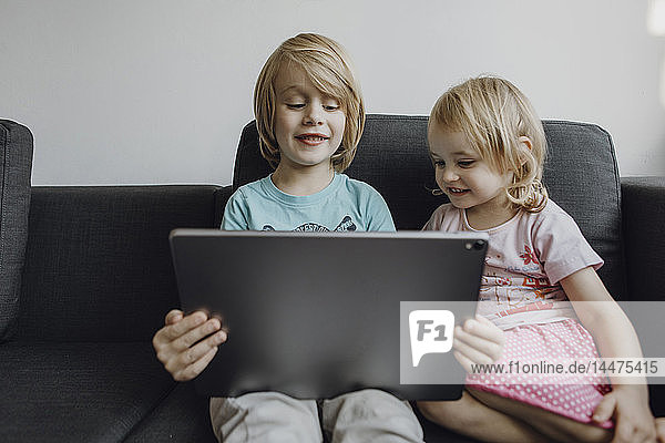 Portrait von Bruder und kleiner Schwester  die zu Hause auf der Couch sitzen und ein digitales Tablet benutzen