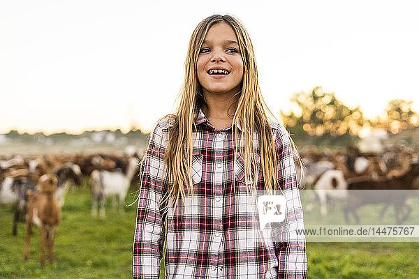 Porträt eines lächelnden blonden Mädchens auf der Weide stehend mit Ziegenherde im Hintergrund
