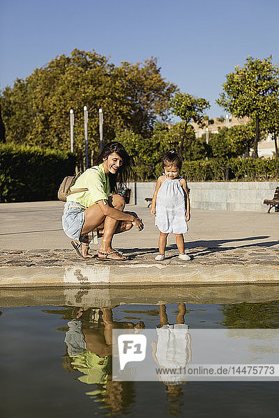 Glückliche Mutter mit Tochter  die ihr Spiegelbild in einem Pool in einem Park betrachten