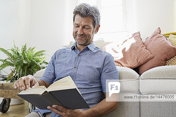 Reifer Mann zu Hause  der vor der Couch sitzt und ein Buch liest
