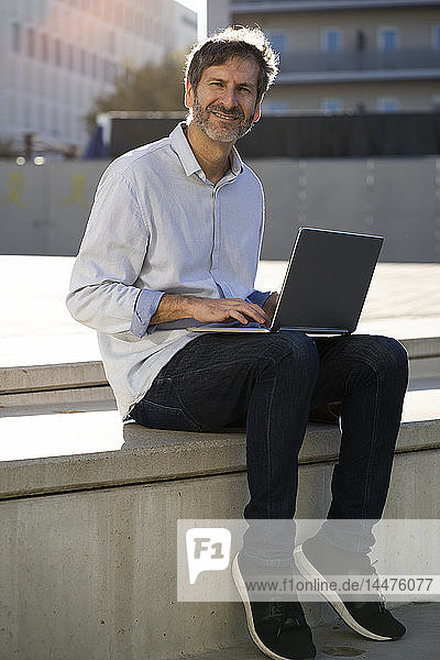 Portrait of smiling mature man sitting in the city using laptop
