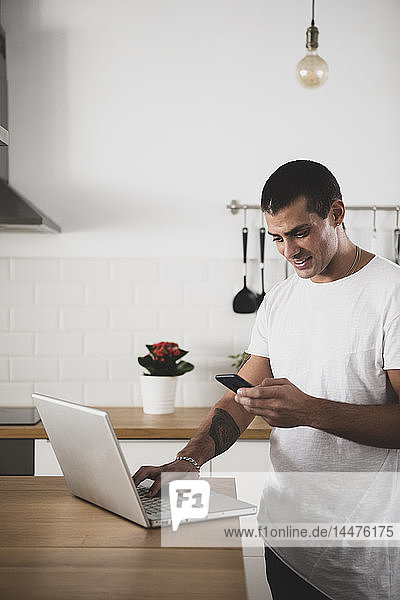Smiling young man using laptop and cell phone in kitchen at home