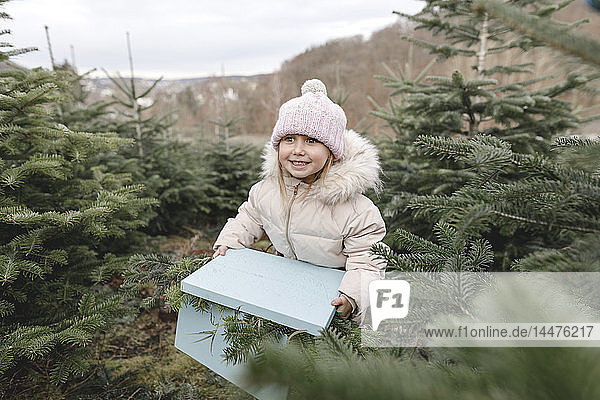 Glückliches Mädchen trägt Geschenkkarton auf einer Weihnachtsbaumplantage