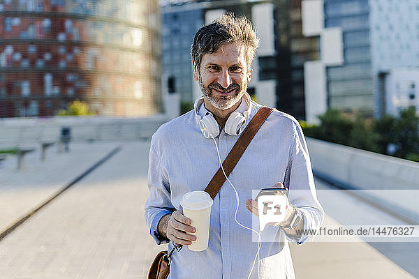 Porträt eines lächelnden reifen Mannes mit Kaffee zum Mitnehmen  Kopfhörer und Mobiltelefon in der Stadt