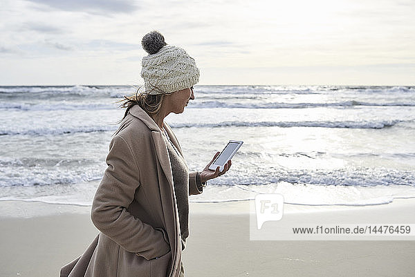 Spain  Menorca  senior woman walking on the beach in winter reading E-Book