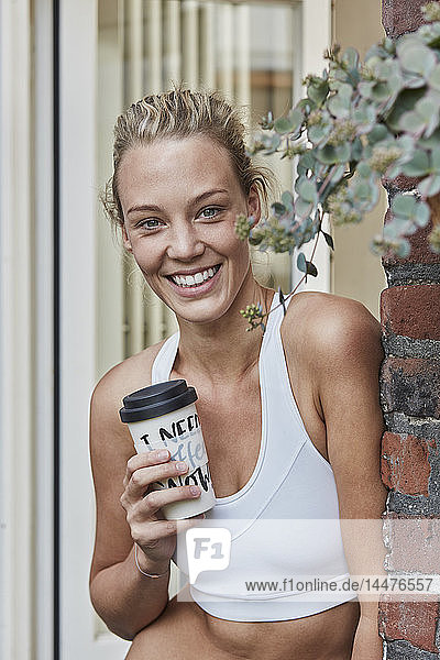 Portrait of smiling sporty young woman with takeaway drink at house entrance