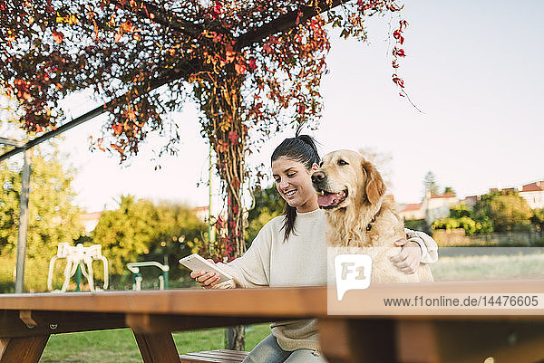 Lächelnde junge Frau mit Handy und ihrem Golden-Retriever-Hund  der sich in einem Park ausruht
