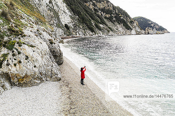 Italien  Elba  Frau mit rotem Mantel beim Fotografieren am Strand  Luftaufnahme mit Drohne