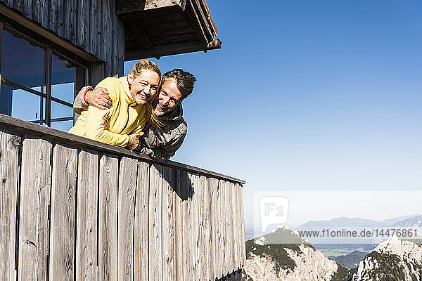 Auf dem Balkon einer Berghütte lehnendes Ehepaar  die Arme um den Kopf gelegt