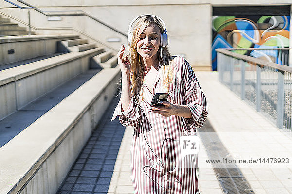 Spain  Barcelona  portrait of smiling young woman using smartphone and headphones outdoors