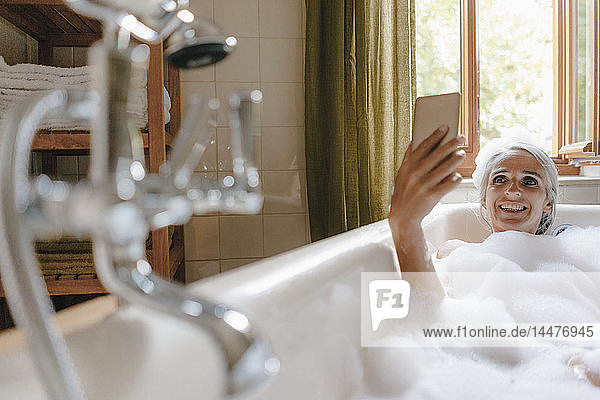Porträt einer glücklichen Frau in der Badewanne  die sich mit dem Handy selbstständig macht