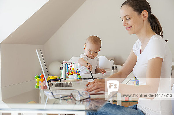 Lächelnde Mutter benutzt Laptop und kleine Tochter spielt zu Hause am Tisch