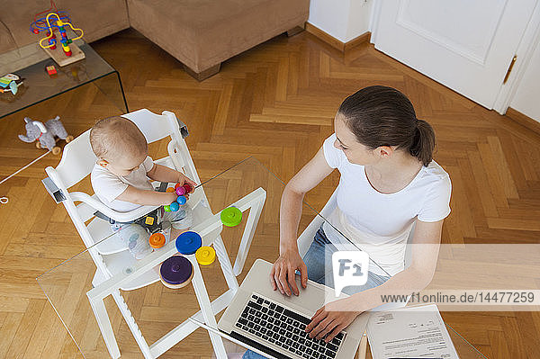 Draufsicht der Mutter am Laptop und der kleinen Tochter beim Spielen am Tisch zu Hause