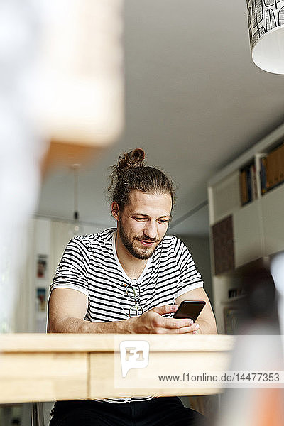 Junger Mann mit Brötchen  der zu Hause sitzt und ein Smartphone benutzt