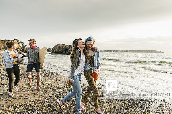 Glückliche Freunde mit Surfbrett und Getränken am steinigen Strand spazieren