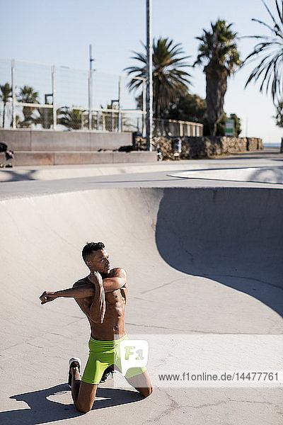 Barechested muscular man doing stretching exercise in a skatepark