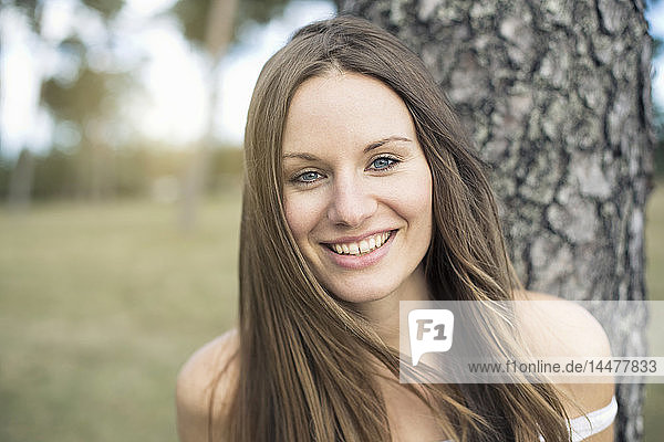 Porträt einer lächelnden jungen Frau  die sich an einen Baumstamm lehnt