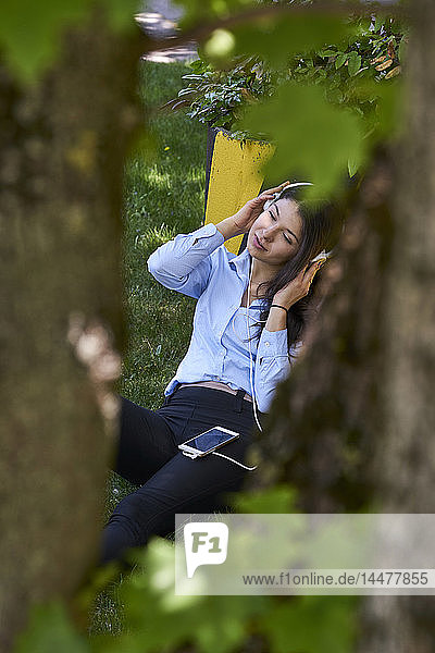 Junge Frau sitzt auf einer Wiese und hört Musik mit Kopfhörern und Handy