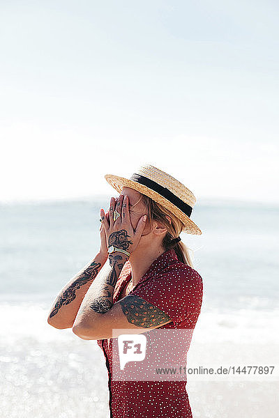 Young woman with tattoos on hand and arms covering her face in front of the sea