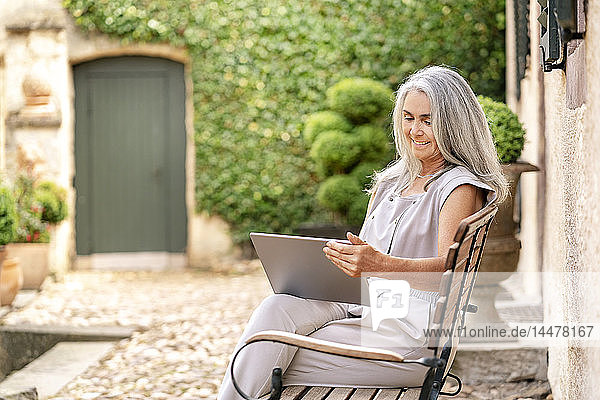 Frau mit langen grauen Haaren sitzt mit Tablette auf Bank im Landhaus