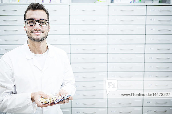 Porträt eines lächelnden Apothekers  der Tablettenpackungen am Schrank in der Apotheke hält