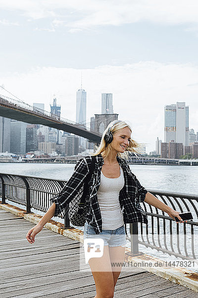 USA  New York City  Brooklyn  glückliche junge Frau mit Kopfhörern und Handy tanzt am Wasser